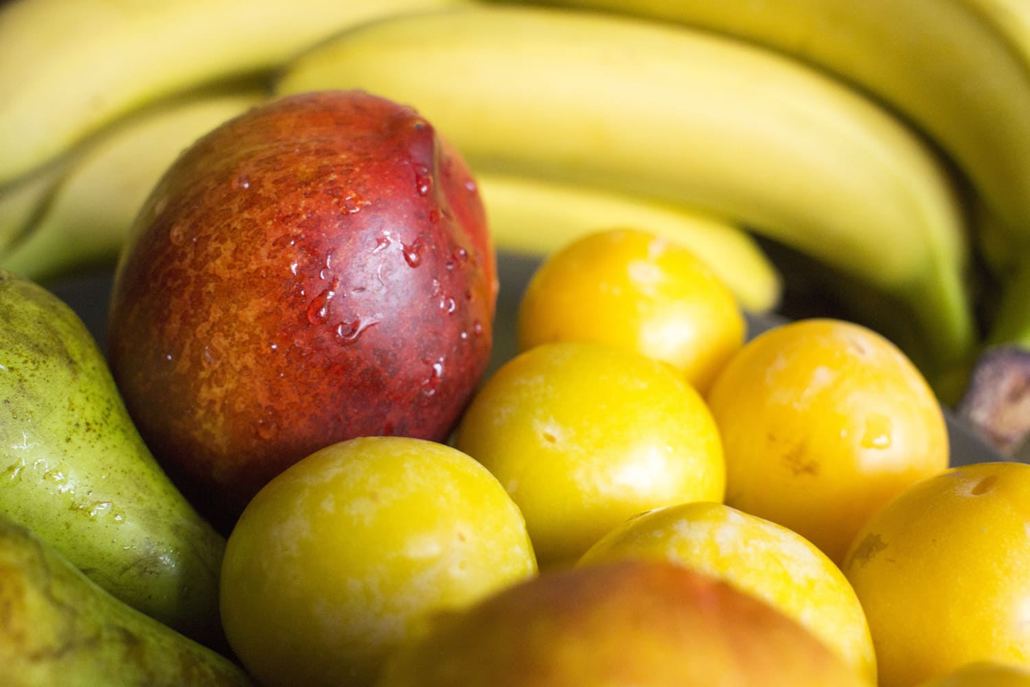 Una selección de frutas frescas incluyendo plátanos, ciruelas amarillas, una pera y una nectarina con gotas de agua.